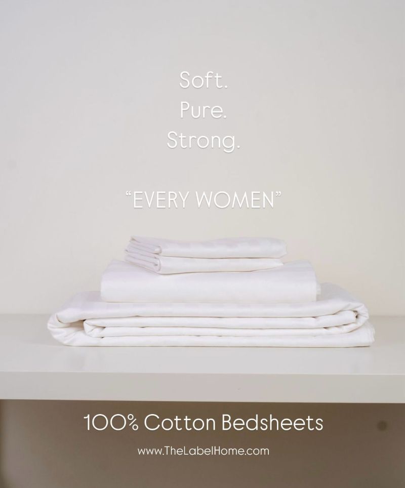 "A neatly folded stack of white 100% cotton bedsheets placed on a shelf, with text overlay reading 'Soft. Pure. Strong. Every Women' and The Label Home website link at the bottom.