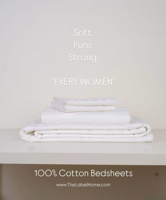 "A neatly folded stack of white 100% cotton bedsheets placed on a shelf, with text overlay reading 'Soft. Pure. Strong. Every Women' and The Label Home website link at the bottom.