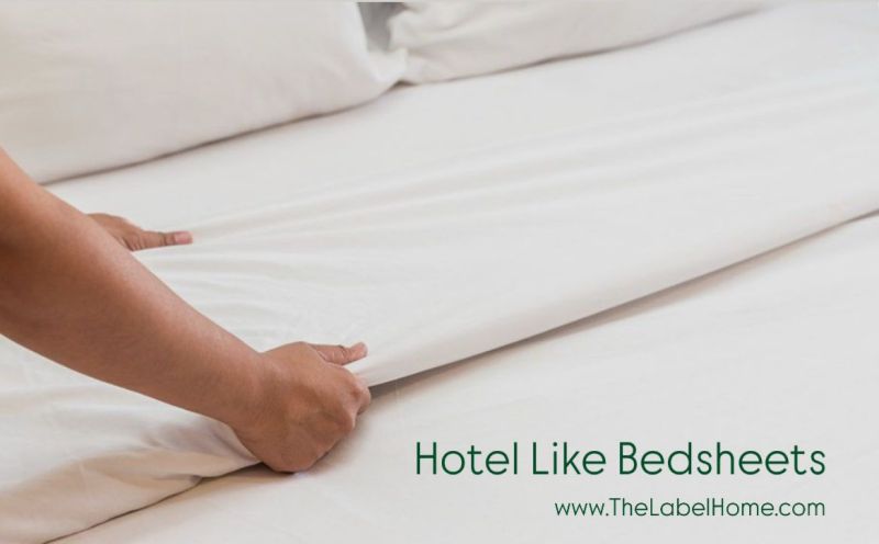 Hands neatly tucking a white bedsheet on a mattress, with text overlay reading 'Hotel Like Bedsheets' and The Label Home website link at the bottom.